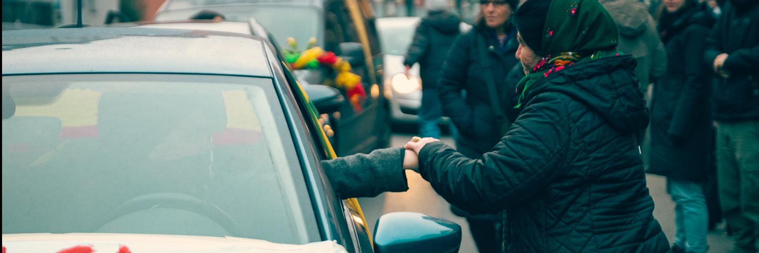 Ein Foto der Karawane: Eine Person streckt die Hand aus dem Fenster eines Autos. Eine Frau ergreift diese und hält sie mit beiden Händen fest. Eine Abschiedsszene.